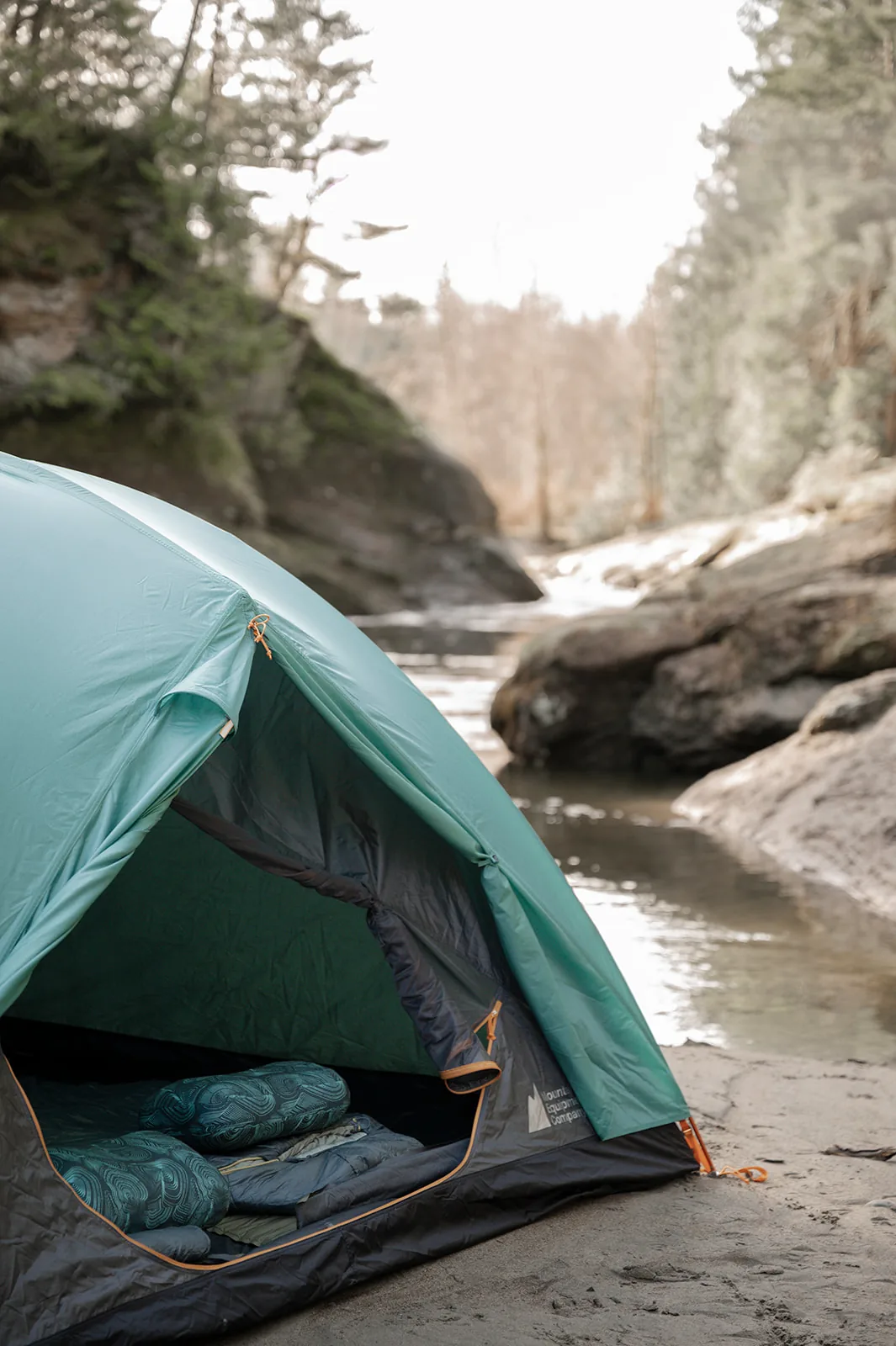 A sage green MEC tent set up on a sandy creekside beach surrounded by forest, sleeping bags visible inside